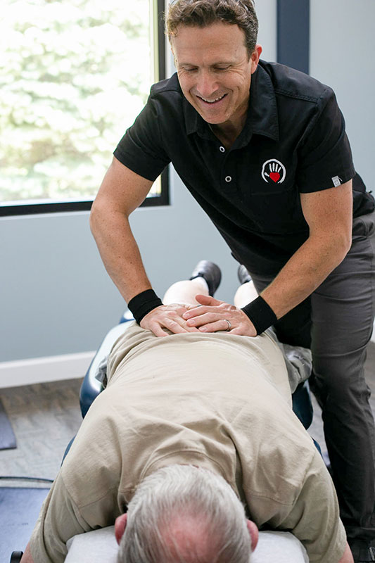 Chiropractor adjusts lower back of old man as he lays face down on adjustment table.