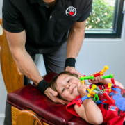 friendly chiropractor adjusts the neck and back of toddler while he lays on his back and play with a toy.